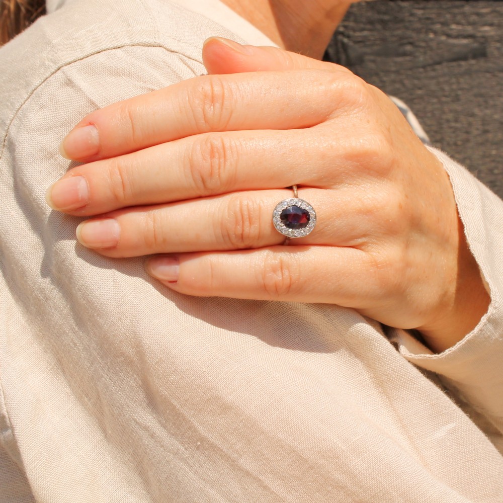 An Art Deco ring featuring an oval deep blue sapphire surrounded by a halo of small diamonds set on a slender silver tone band. View VI