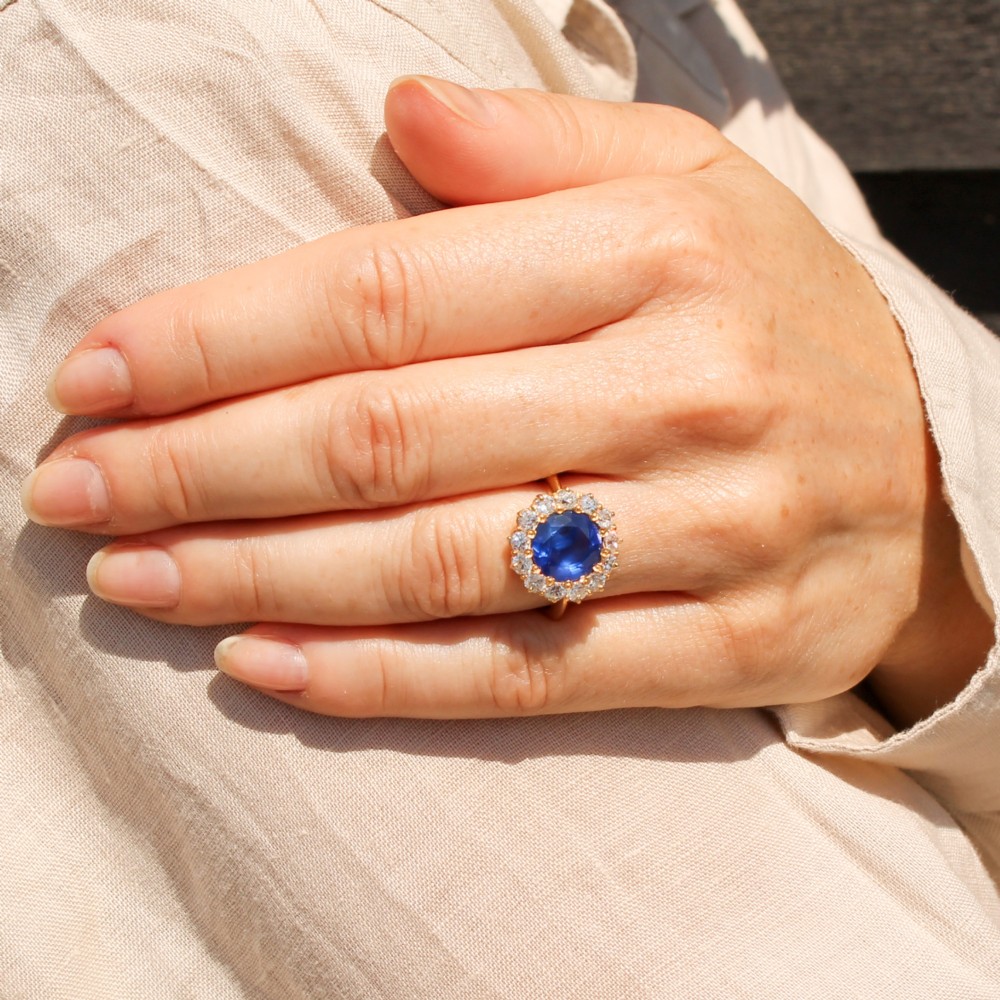 An Edwardian cluster ring set with a circular blue central sapphire framed by a halo of old cut diamonds on a yellow gold band. View II
