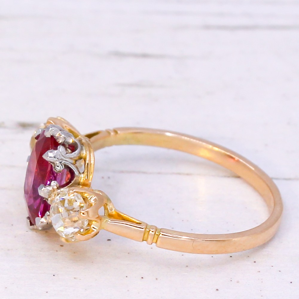 An antique yellow gold ring set with a central oval pinkish-red ruby flanked by two round old cut diamonds in claw settings. View IV
