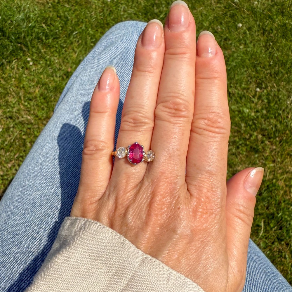 An antique yellow gold ring set with a central oval pinkish-red ruby flanked by two round old cut diamonds in claw settings. View VI