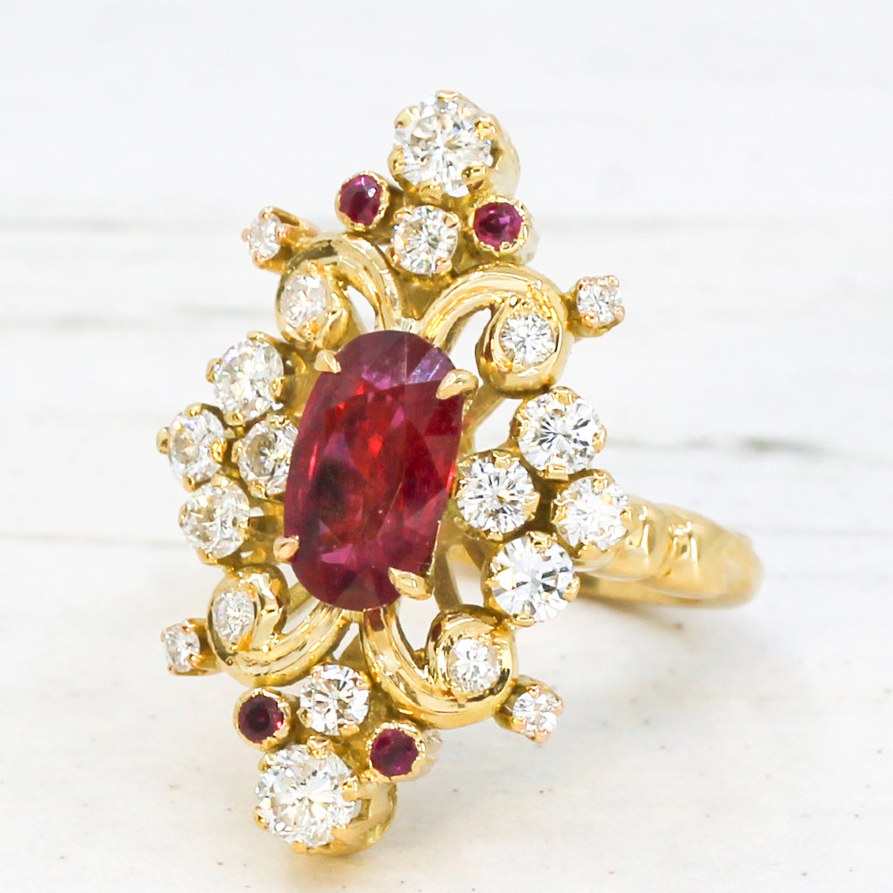 A yellow gold vintage ring featuring an oval red ruby surrounded by openwork scroll motifs set with round diamonds and accent rubies.