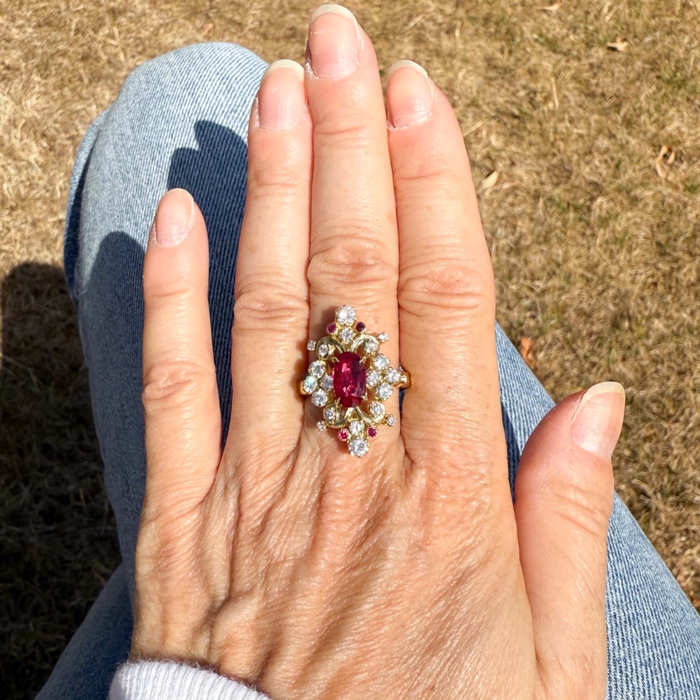 A yellow gold vintage ring featuring an oval red ruby surrounded by openwork scroll motifs set with round diamonds and accent rubies. View VI