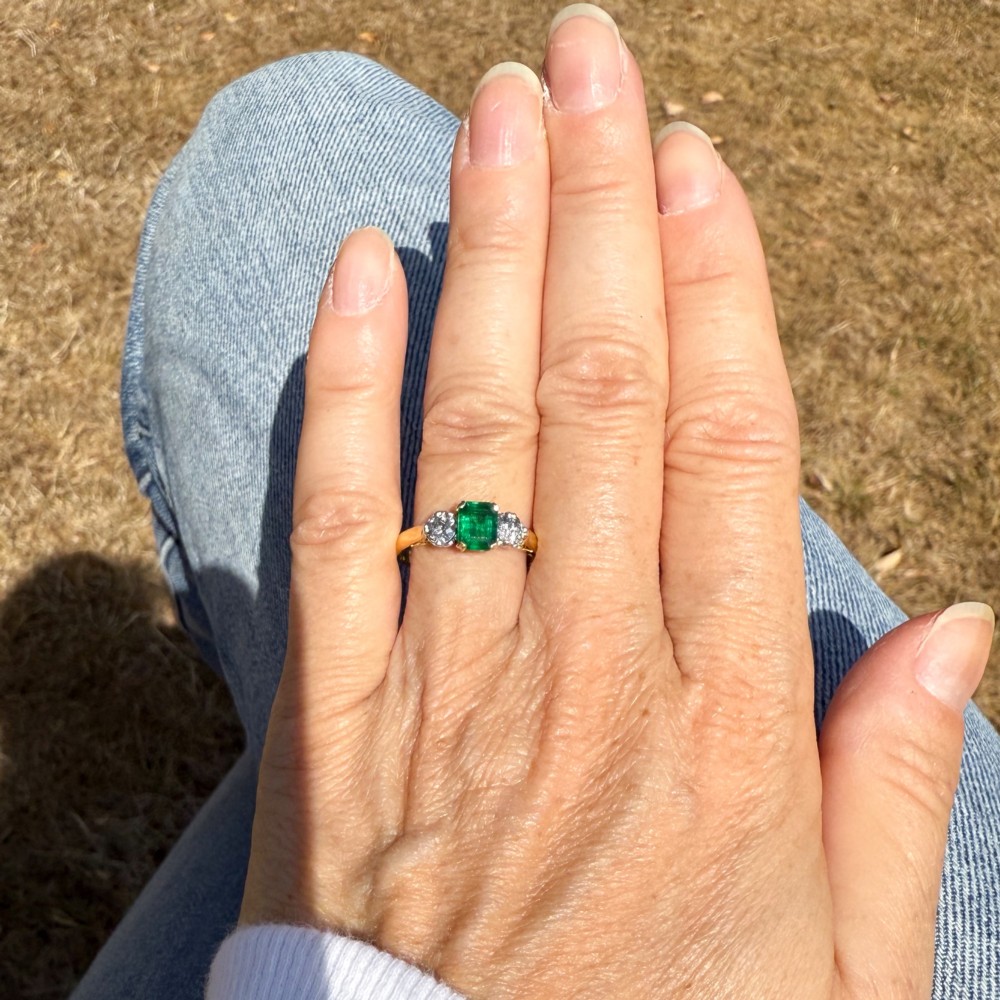 A yellow gold band holding a central rectangular green emerald flanked by two round clear diamonds in claw settings. View VI