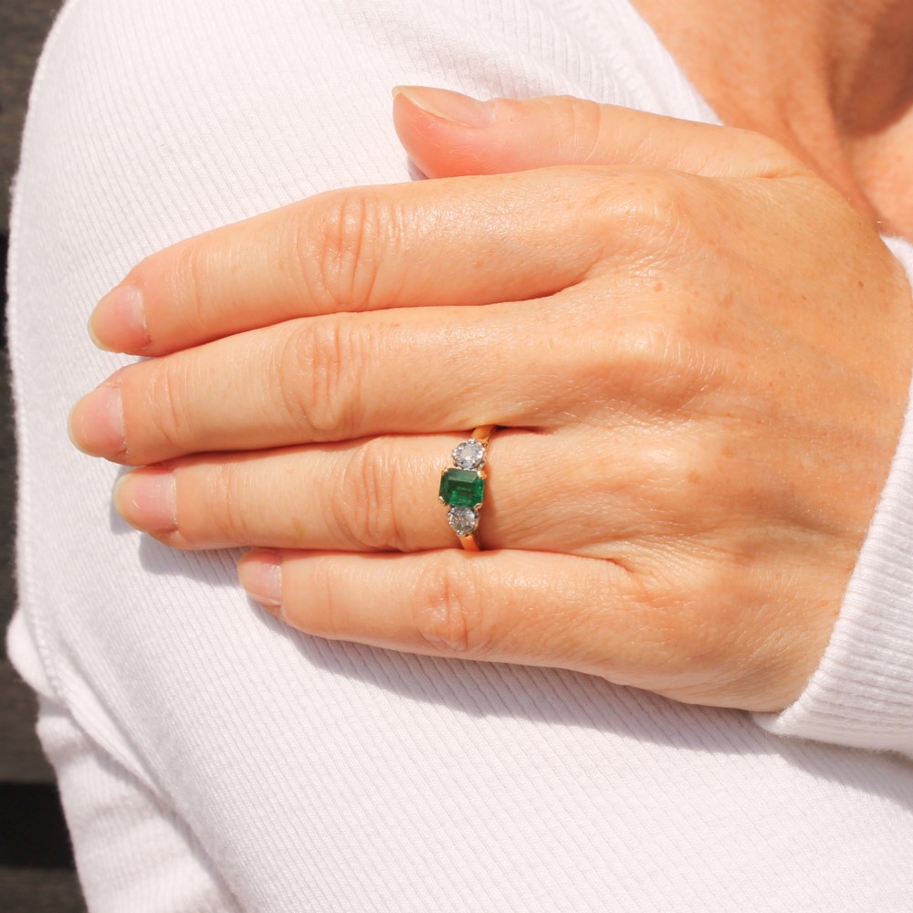 A yellow gold band holding a central rectangular green emerald flanked by two round clear diamonds in claw settings. View VII