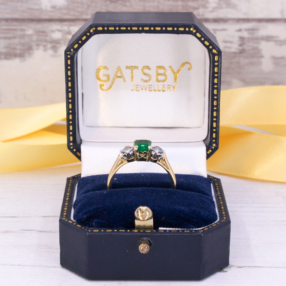 A yellow gold band holding a central rectangular green emerald flanked by two round clear diamonds in claw settings. View IX