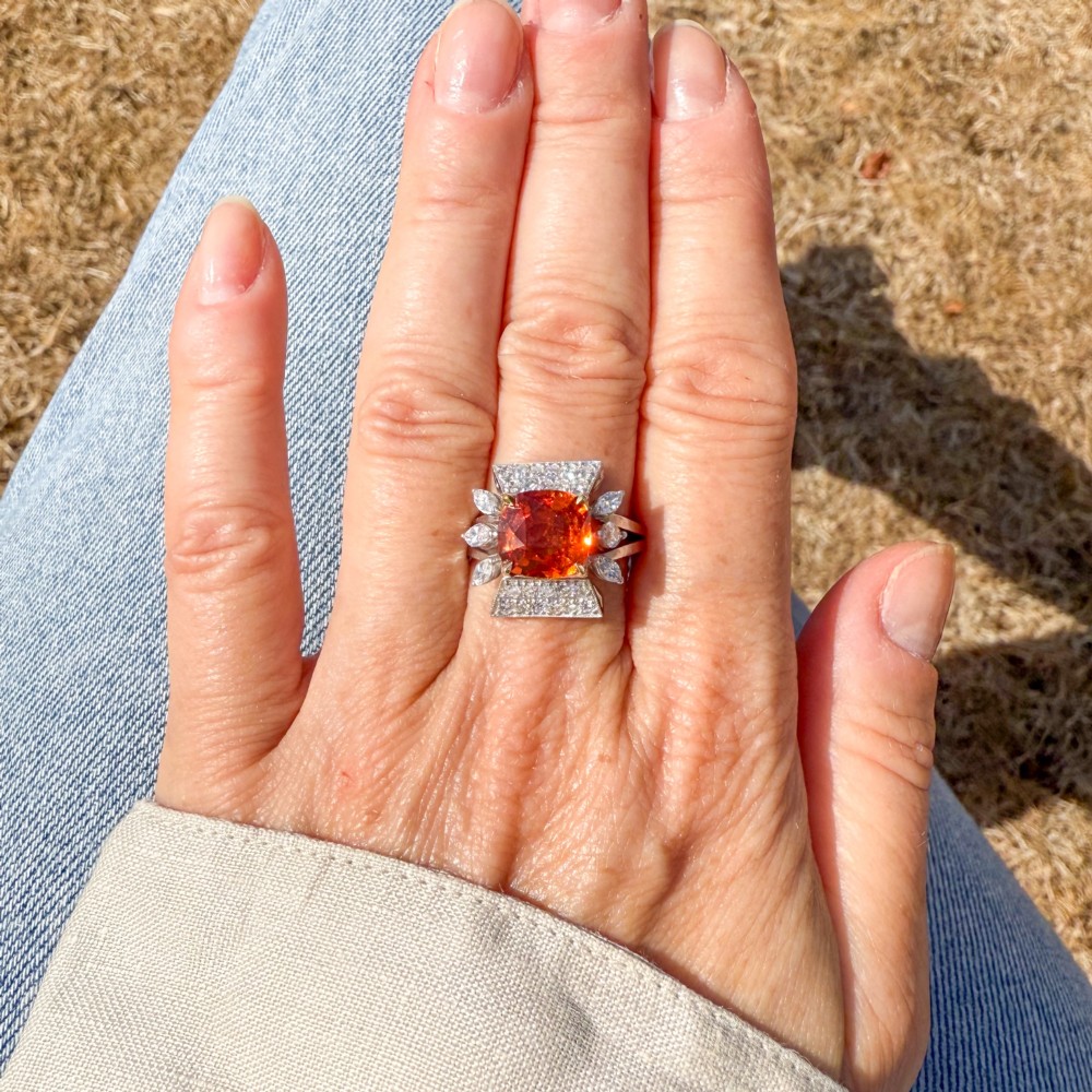 A mid-century vintage cocktail ring featuring a cushion-cut orange garnet central gemstone flanked by pave and marquise diamond accents set in a white gold band. View VI
