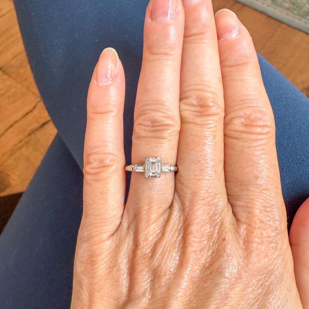 A silver-toned metal ring featuring a central rectangular emerald-cut diamond flanked by two baguette-cut diamonds on the shoulders. View VII