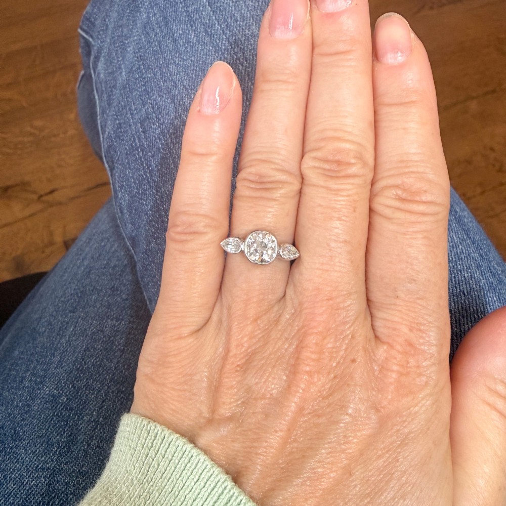 An Art Deco platinum ring featuring a central bezel-set old cushion-cut diamond flanked by two pear-cut side diamonds with milgrain edging. View VI