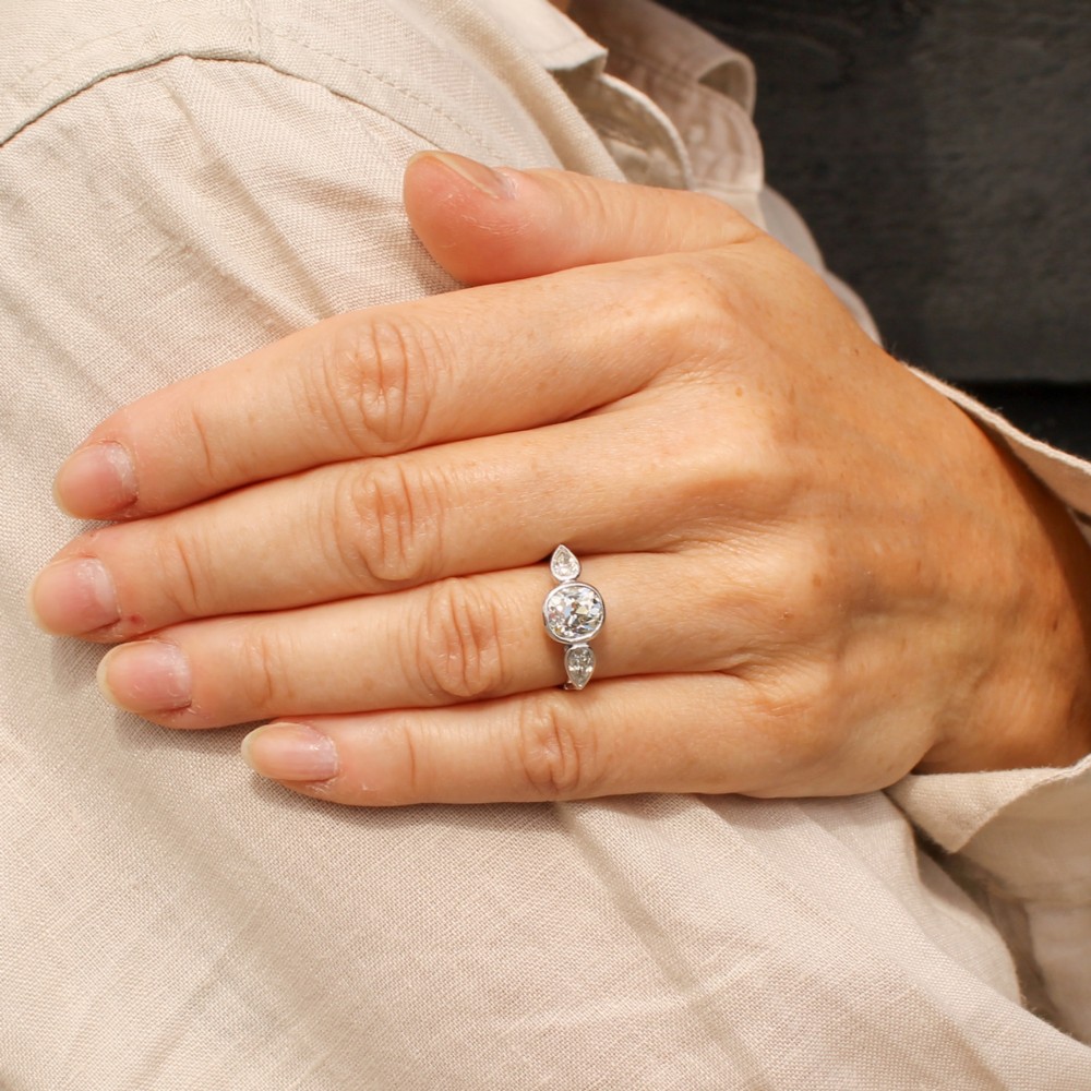 An Art Deco platinum ring featuring a central bezel-set old cushion-cut diamond flanked by two pear-cut side diamonds with milgrain edging. View VII