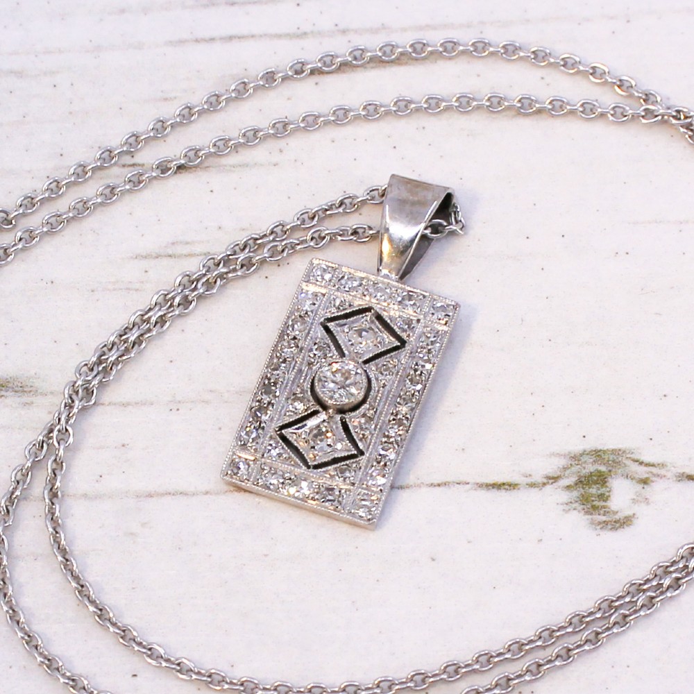 A rectangular Art Deco pendant set with a central round old cut diamond within a geometric diamond-paved frame on a silver chain.