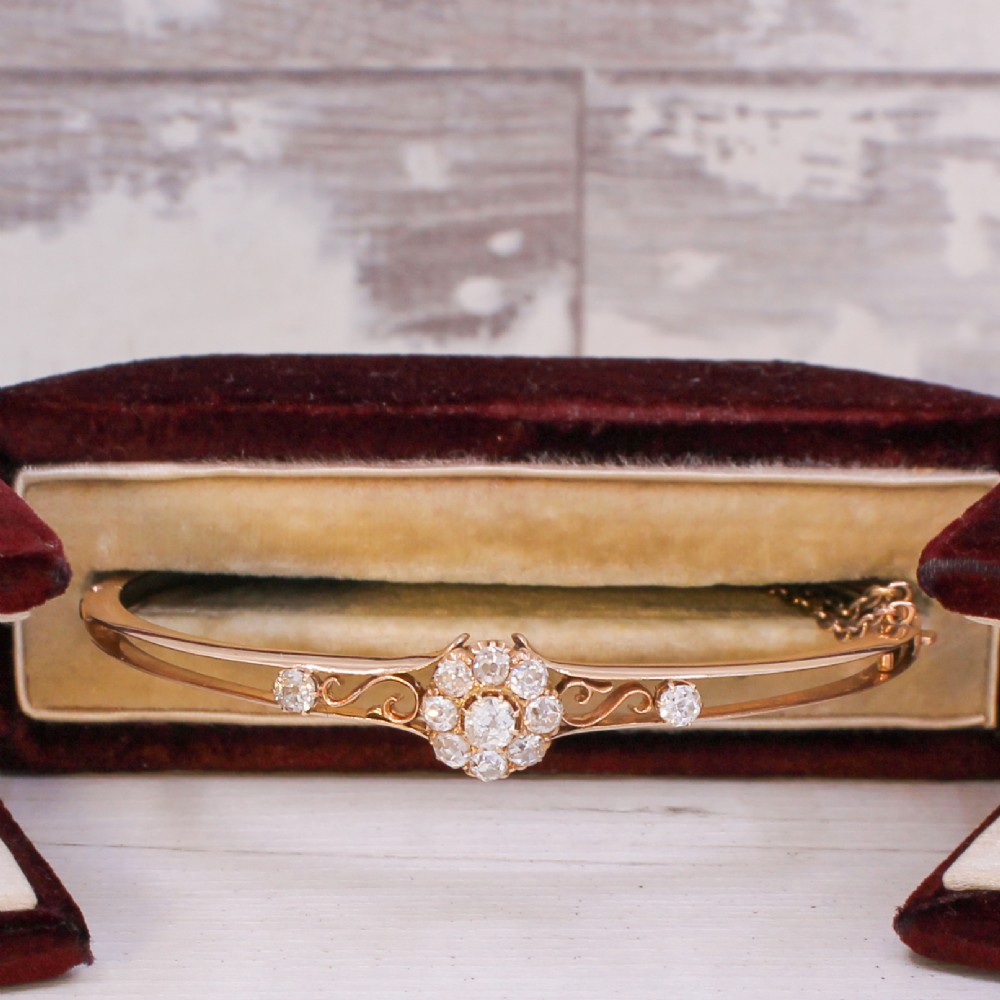 A late Victorian yellow gold hinged bangle featuring a central old-cut diamond cluster flanked by scrollwork and accent diamonds. View X