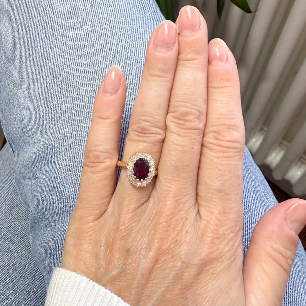 A vintage yellow gold ring featuring a central oval-cut red ruby framed by a halo of brilliant round diamonds. View VI