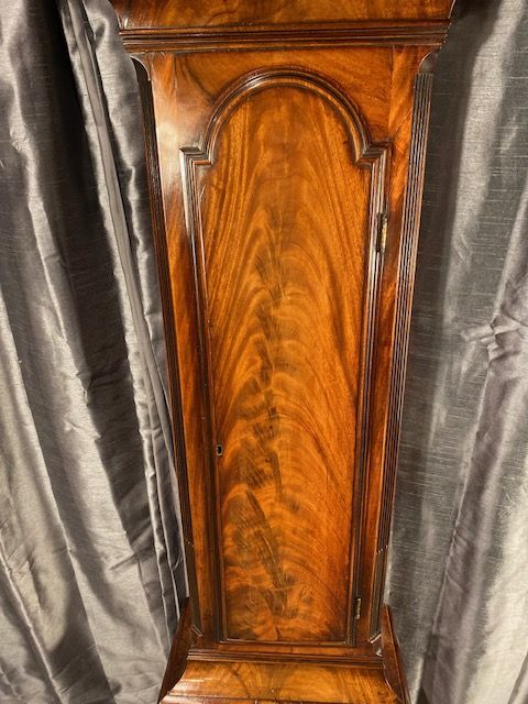 fig 2. A tall antique flame mahogany longcase clock featuring an arched dial with Roman numerals signed James McCabe London and topped with a brass finial.