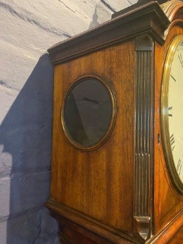 fig 1. A tall flame mahogany longcase clock with an arched hood, brass finial, and round dial face.