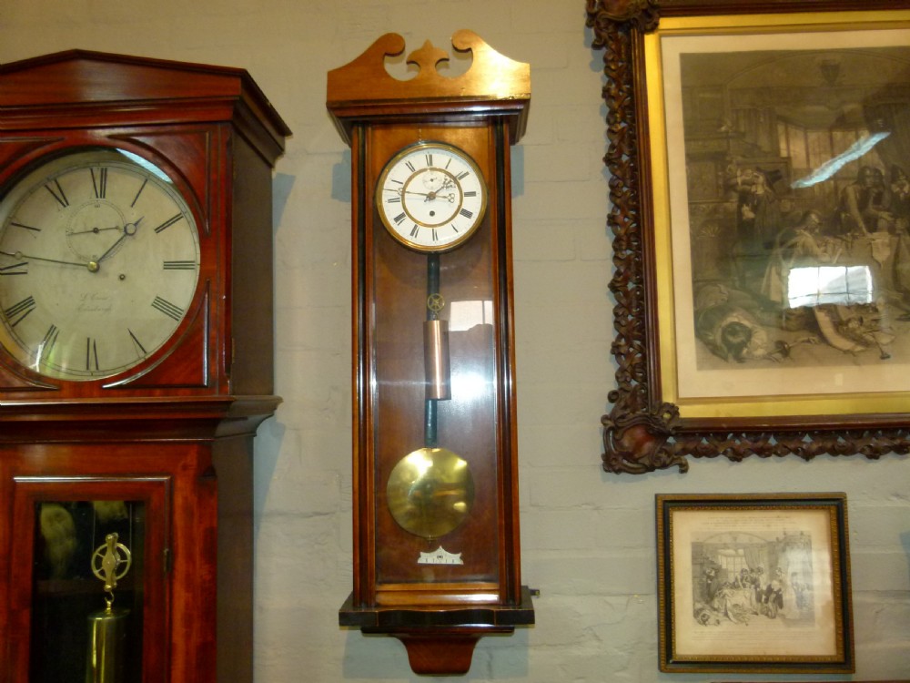 A mahogany wall-mounted regulator clock with a scroll pediment, white dial with Roman numerals, and visible brass weight and pendulum within a glazed door.