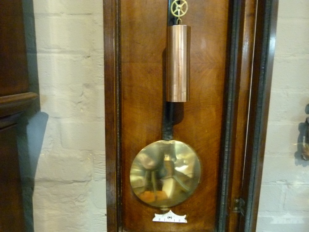 A mahogany wall-mounted regulator clock with a scroll pediment, white dial with Roman numerals, and visible brass weight and pendulum within a glazed door. View IV