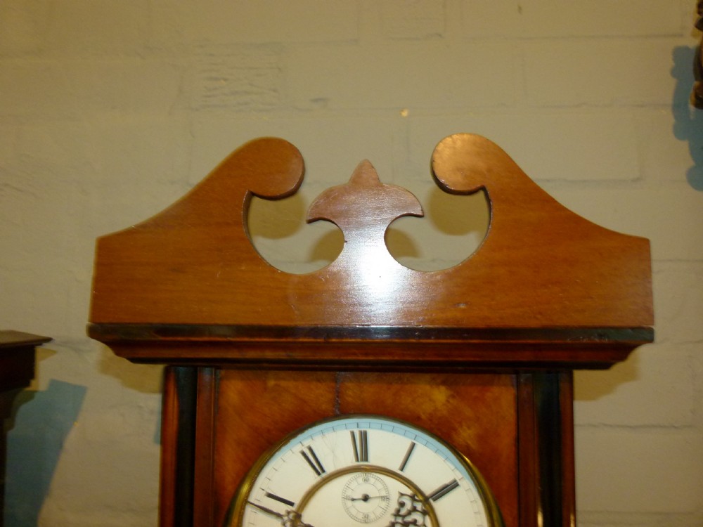 A mahogany wall-mounted regulator clock with a scroll pediment, white dial with Roman numerals, and visible brass weight and pendulum within a glazed door. View V