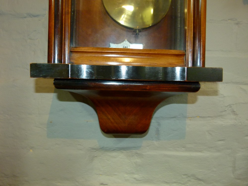 A mahogany wall-mounted regulator clock with a scroll pediment, white dial with Roman numerals, and visible brass weight and pendulum within a glazed door. View VI
