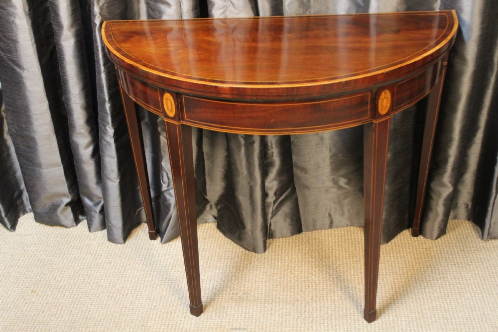 A semi-circular antique mahogany demilune card table featuring satinwood stringing inlays and slender tapered legs.