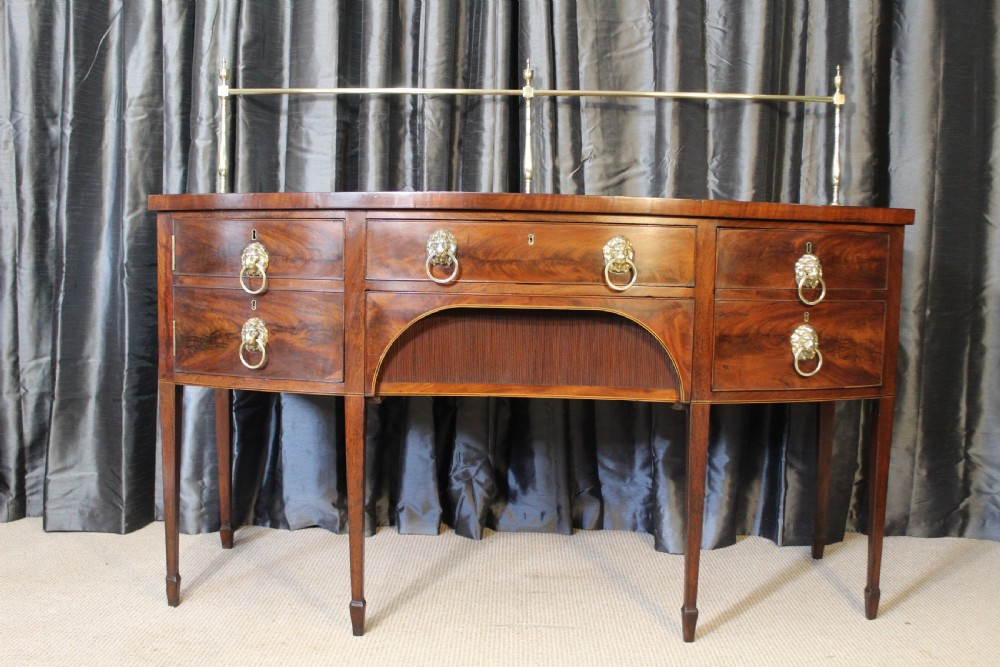 A mahogany bowfront sideboard featuring lion mask brass ring handles, boxwood stringing inlay, square tapered legs with spade feet, and an upper brass rail gallery.