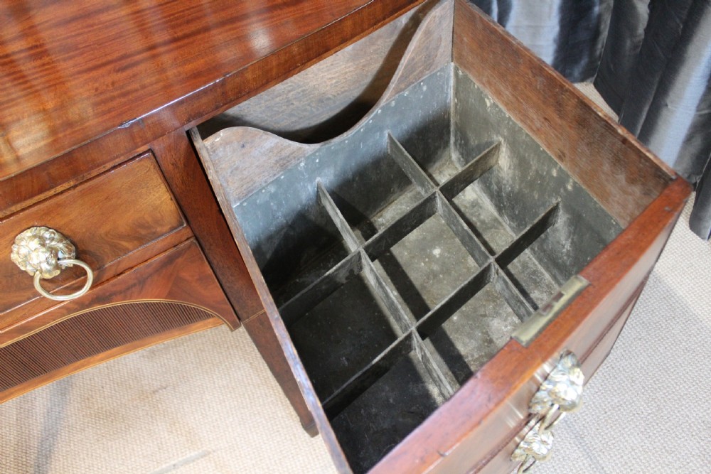 A mahogany bowfront sideboard featuring lion mask brass ring handles, boxwood stringing inlay, square tapered legs with spade feet, and an upper brass rail gallery. View IV