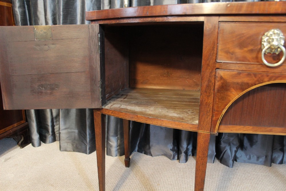 A mahogany bowfront sideboard featuring lion mask brass ring handles, boxwood stringing inlay, square tapered legs with spade feet, and an upper brass rail gallery. View VI