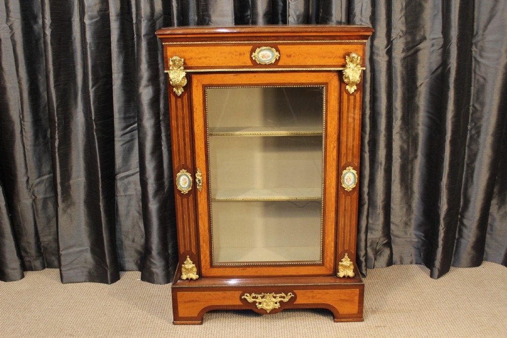 An antique warm brown timber cabinet featuring a glazed front door, ornamental gilt metal mounts, and porcelain floral plaques.