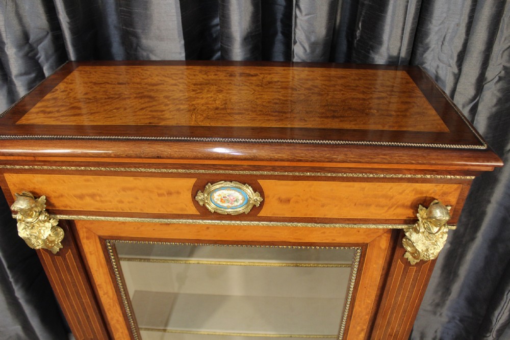 An antique warm brown timber cabinet featuring a glazed front door, ornamental gilt metal mounts, and porcelain floral plaques. View II