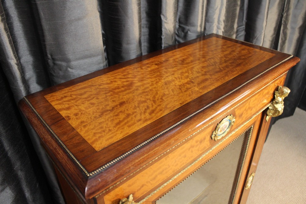 An antique warm brown timber cabinet featuring a glazed front door, ornamental gilt metal mounts, and porcelain floral plaques. View III