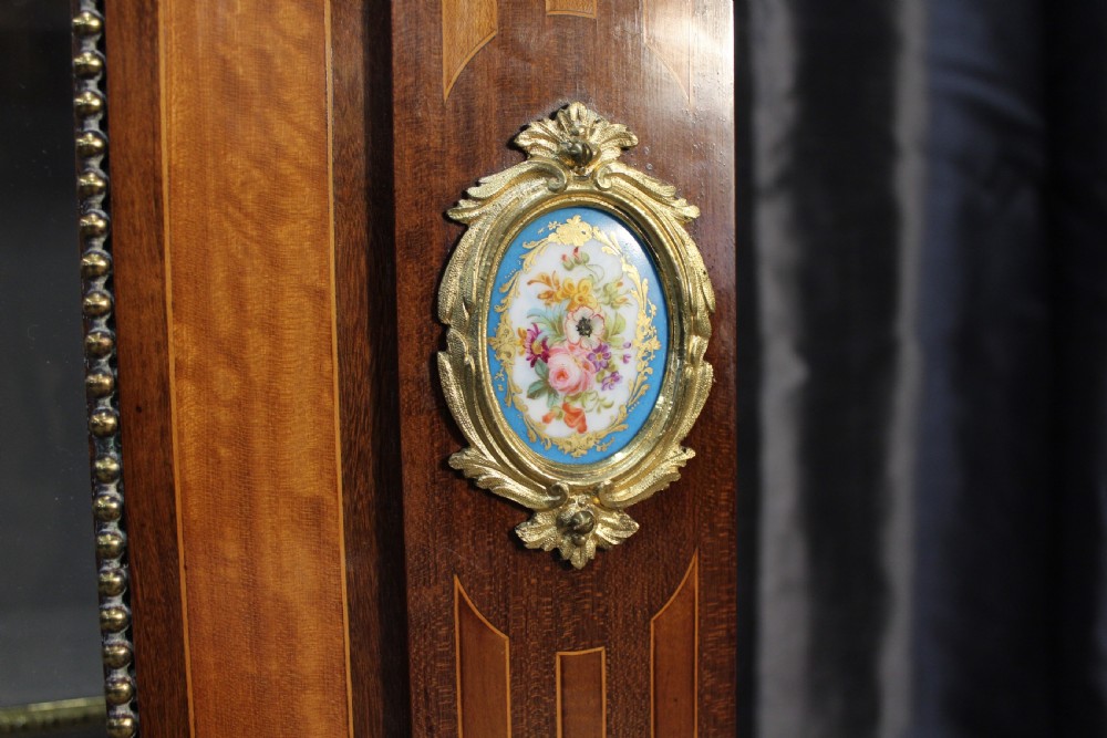 An antique warm brown timber cabinet featuring a glazed front door, ornamental gilt metal mounts, and porcelain floral plaques. View V
