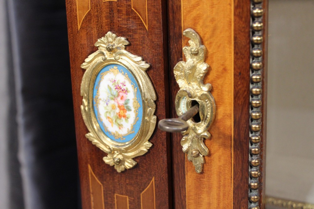 An antique warm brown timber cabinet featuring a glazed front door, ornamental gilt metal mounts, and porcelain floral plaques. View VI