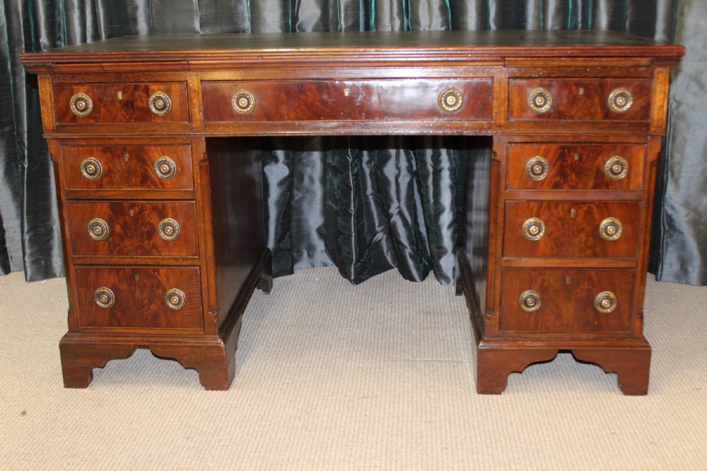 An antique mahogany pedestal desk with nine drawers, circular brass ring handles, flame mahogany drawer fronts, and bracket feet.
