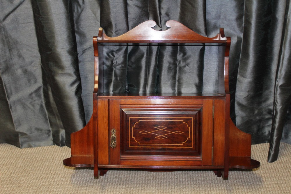 An antique mahogany wall hanging cabinet featuring a central cupboard door with geometric boxwood inlay, a single brass drop handle, a shaped top gallery shelf, and lower side brackets.