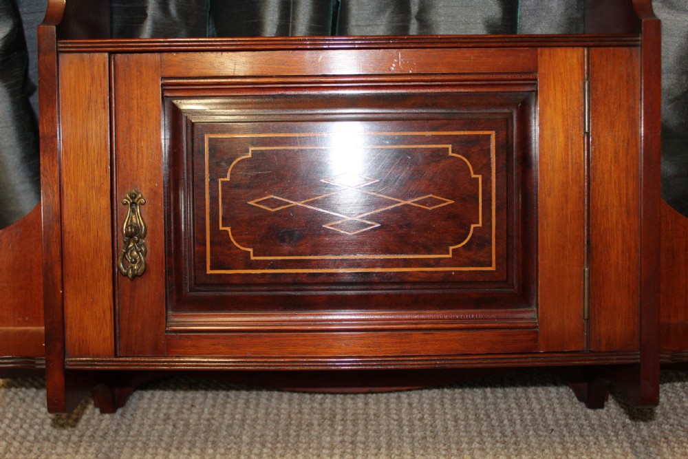 An antique mahogany wall hanging cabinet featuring a central cupboard door with geometric boxwood inlay, a single brass drop handle, a shaped top gallery shelf, and lower side brackets. View II