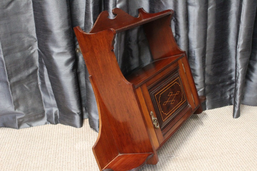 An antique mahogany wall hanging cabinet featuring a central cupboard door with geometric boxwood inlay, a single brass drop handle, a shaped top gallery shelf, and lower side brackets. View V