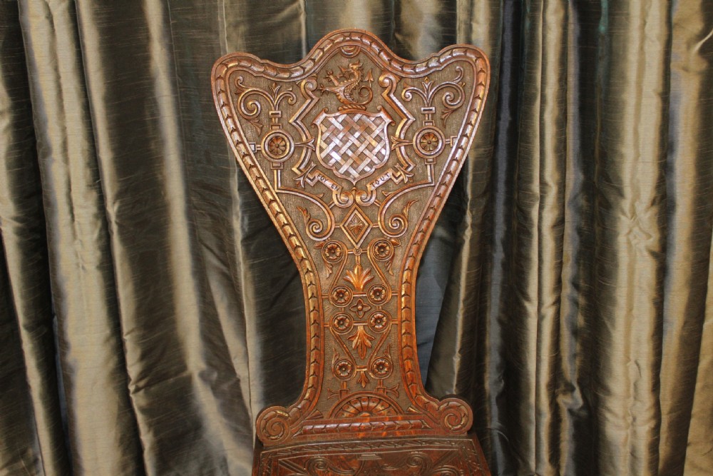 A pair of dark carved oak hall chairs featuring tall shaped backrests with heraldic crest motifs and decorative carved slab bases. View II