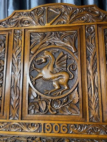 An Edwardian carved oak hall bench dated 1906, featuring mythological beast motifs, foliage panels, carved beast head armrests, and two lower drawers. View VI