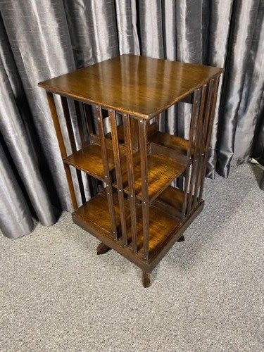 An antique dark oak revolving bookcase featuring a square top, vertical slatted sides, three open tiers, and a four-footed rotating base.