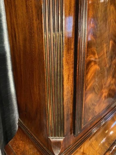 A tall flame mahogany longcase clock with an arched hood, brass finial, and round dial face. View IV
