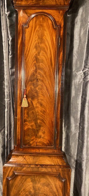 A tall flame mahogany longcase clock with an arched hood, brass finial, and round dial face. View V