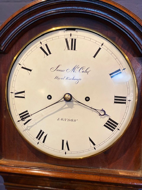 A tall flame mahogany longcase clock with an arched hood, brass finial, and round dial face. View VI