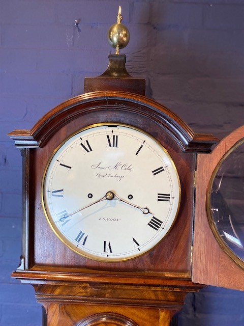 A tall flame mahogany longcase clock with an arched hood, brass finial, and round dial face. View VIII