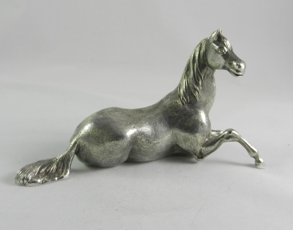 A cast silver figurine depicting a reclining horse with a raised front leg and textured mane.