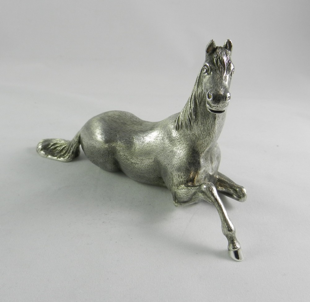 A cast silver figurine depicting a reclining horse with a raised front leg and textured mane. View II