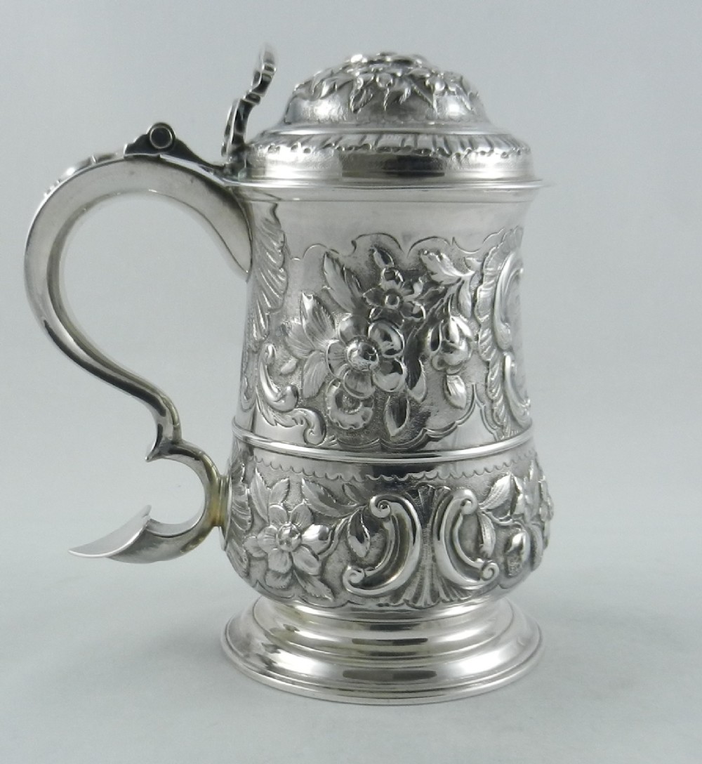 An 18th-century Georgian sterling silver lidded tankard with a scroll handle, hinged dome lid, and intricate floral repousse chasing across the body.