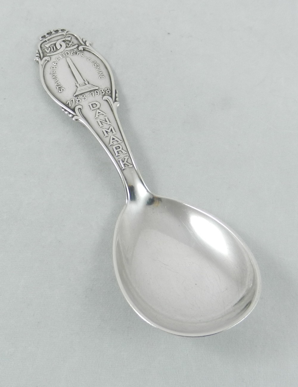 A silver Danish caddy spoon featuring a shaped bowl and an embossed handle depicting an obelisk motif and Danish lettering.