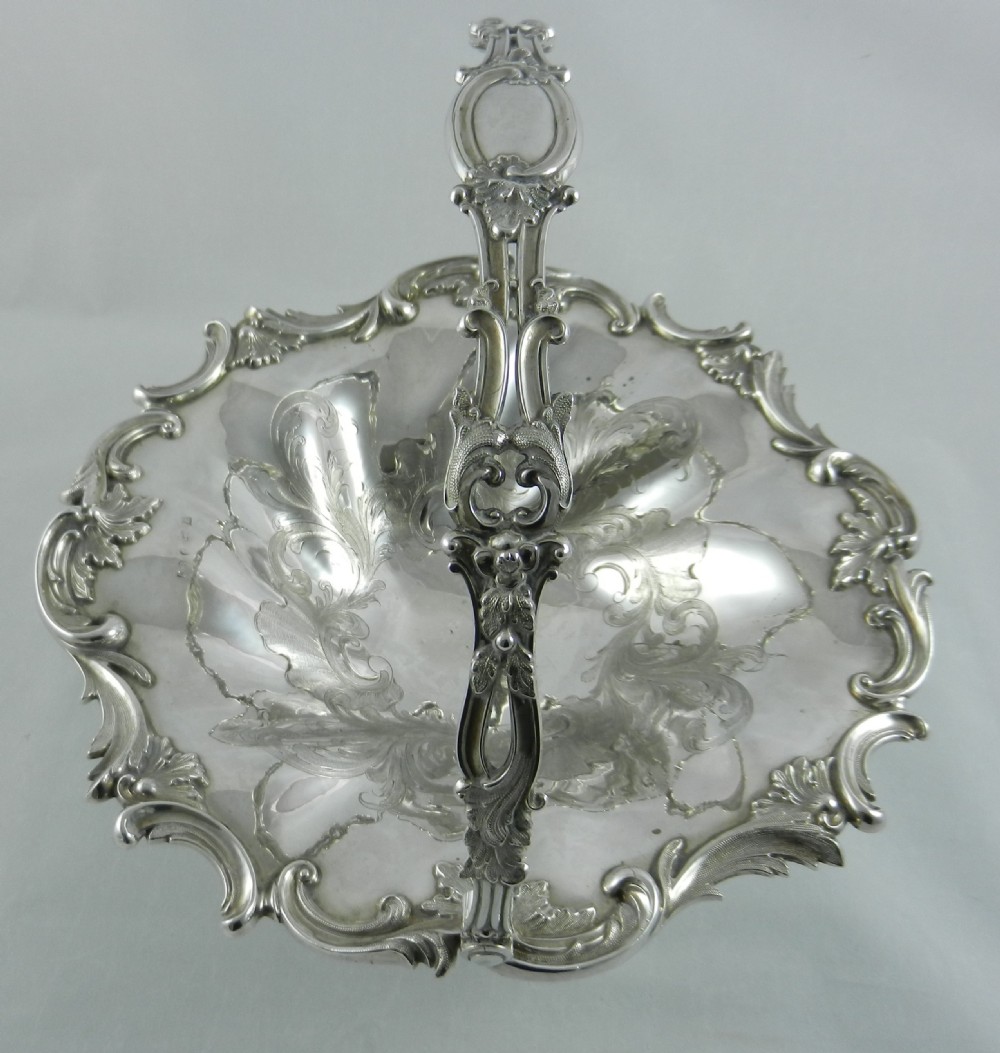 An antique silver cake basket featuring a swing handle, floral engraved interior, scrolled rim, and a pedestal base. View III
