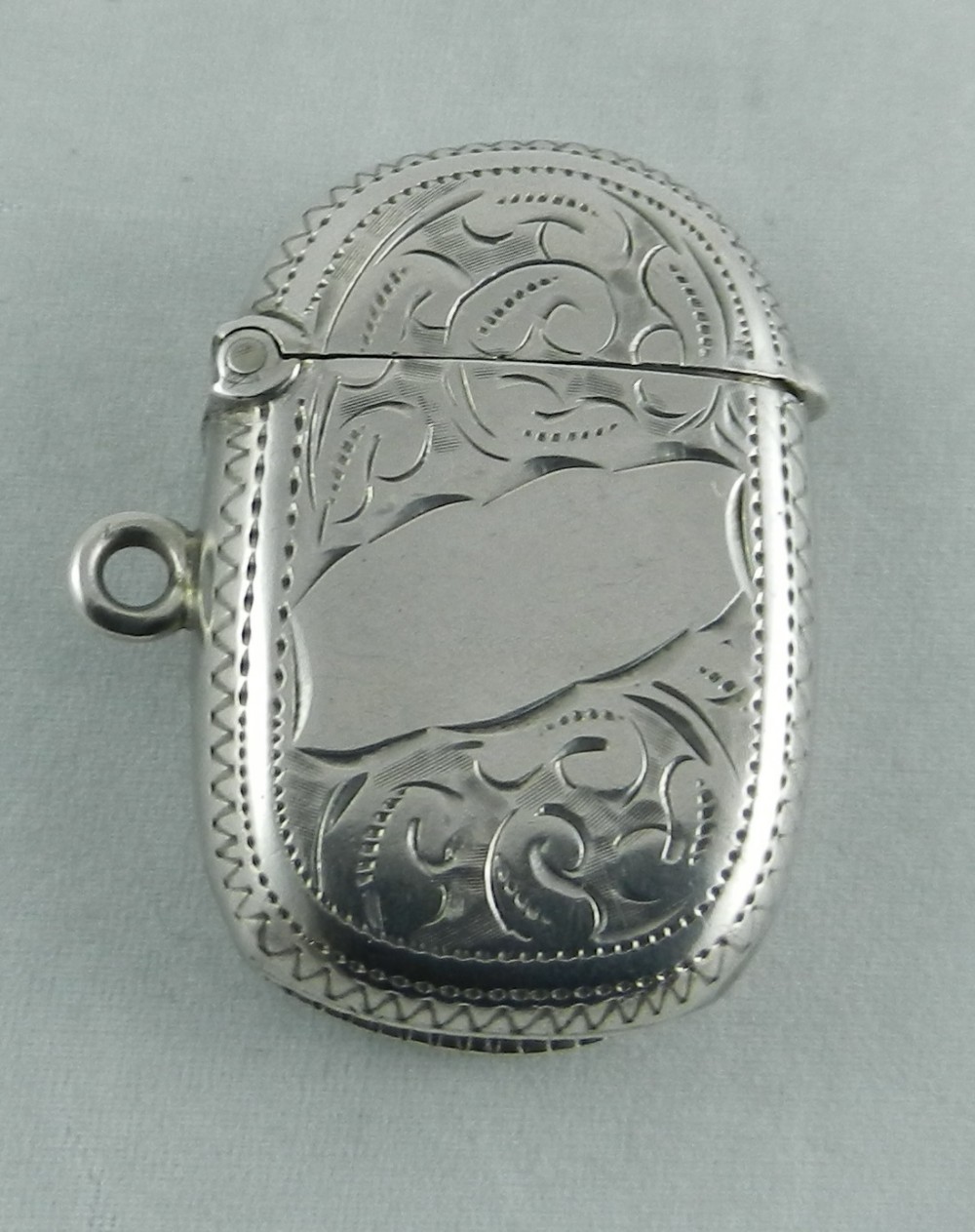 An oval sterling silver vesta case featuring hand-engraved foliate scrollwork, a blank central cartouche, a hinged lid, and a side suspension ring.