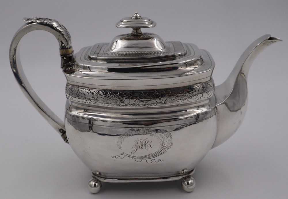 An antique sterling silver teapot with engraved foliate motifs, an oblong body, ball feet, and a hinged lid.