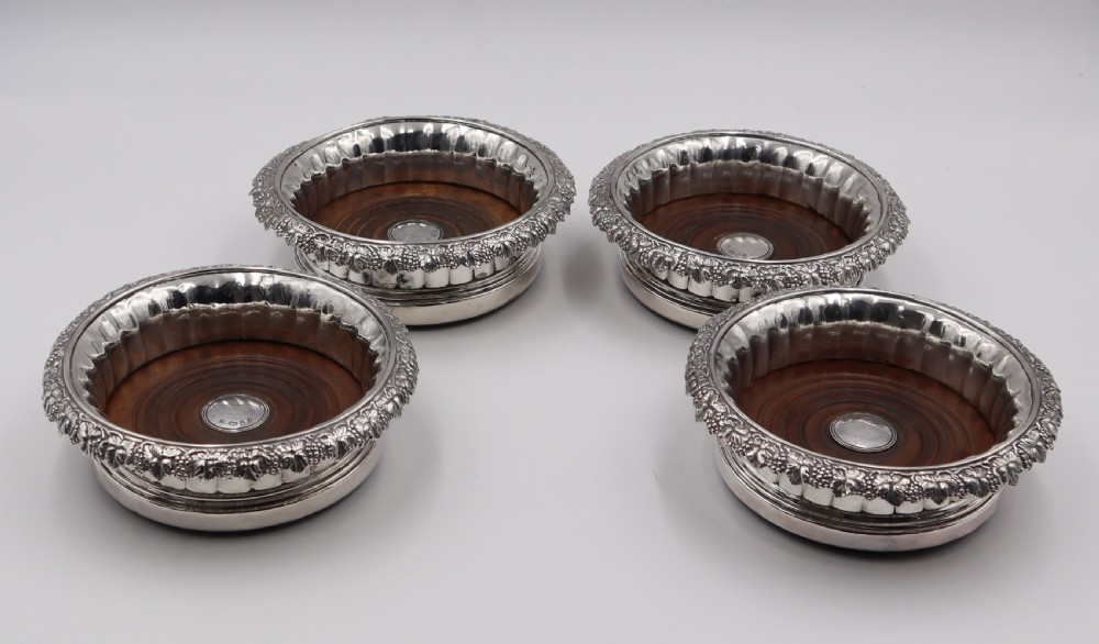 A set of four circular silver wine coasters featuring fluted inner rims, embossed grape and vine borders, and turned wood bases with central silver bosses.